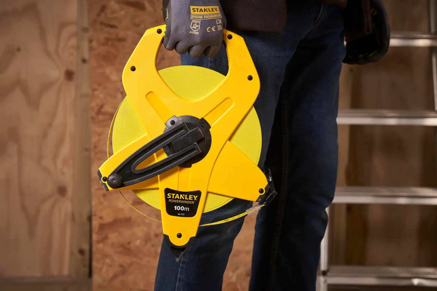 STANLEY 2-34-777 metro a nastro con avvolgitore aperto tenuto da persona con guanti, sfondo da cantiere.