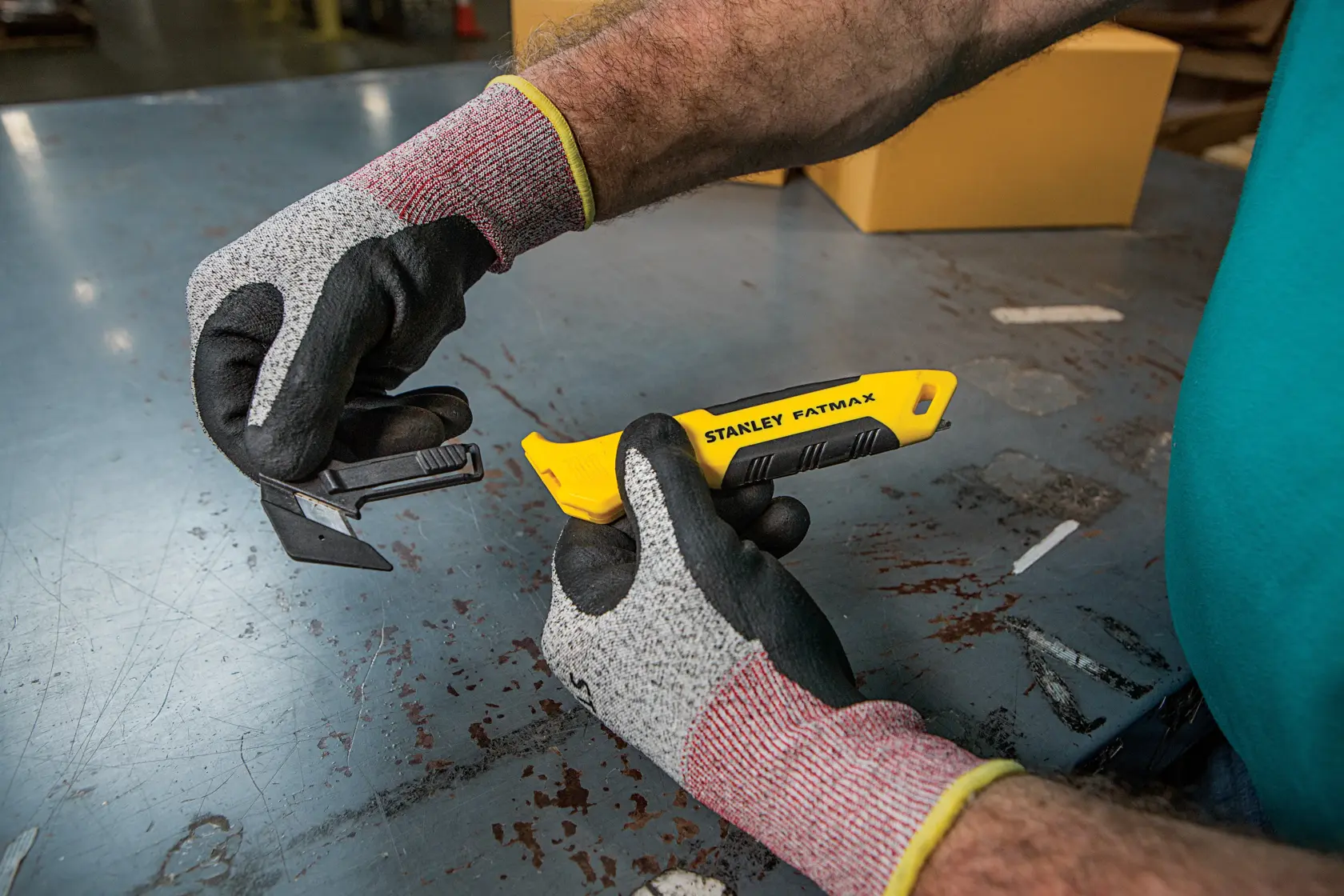 Foto di taglierino STANLEY FMHT10358-0 con manico giallo tenuto da mani guantate su tavolo di metallo.
