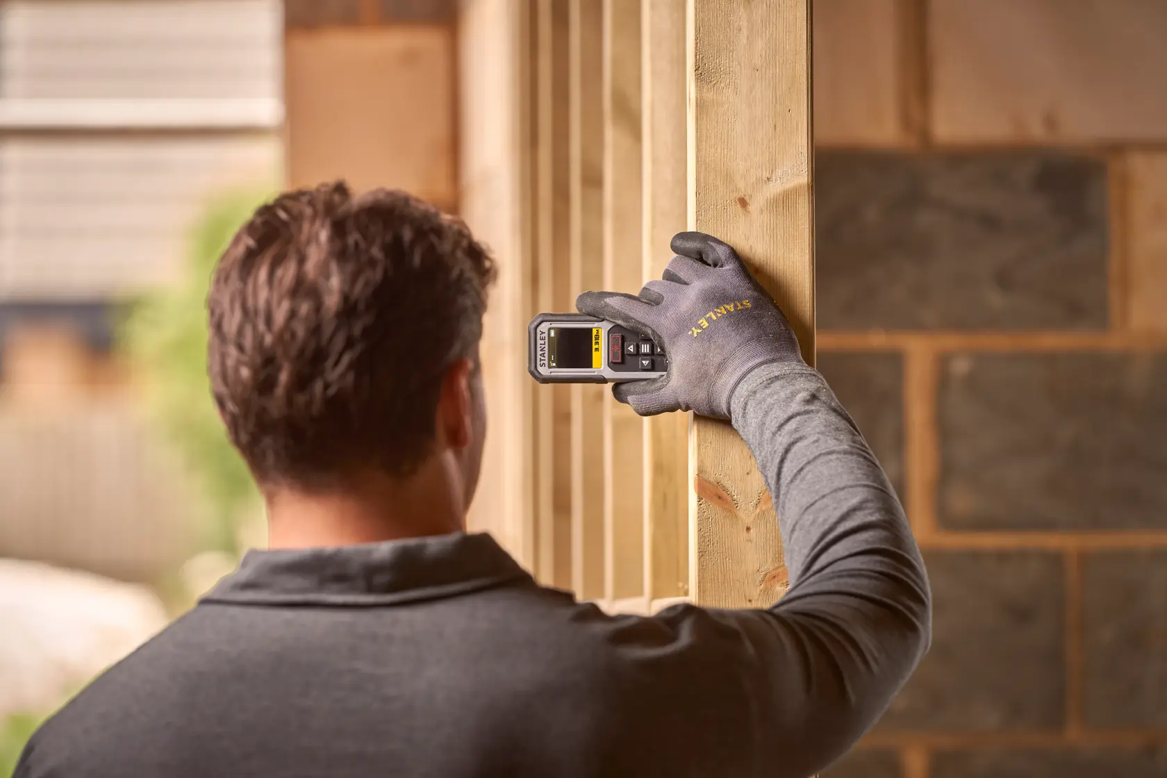 Misuratore laser STANLEY FMHT77050-XJ tenuto da mano con guanto su trave in legno, sfondo cantiere.
