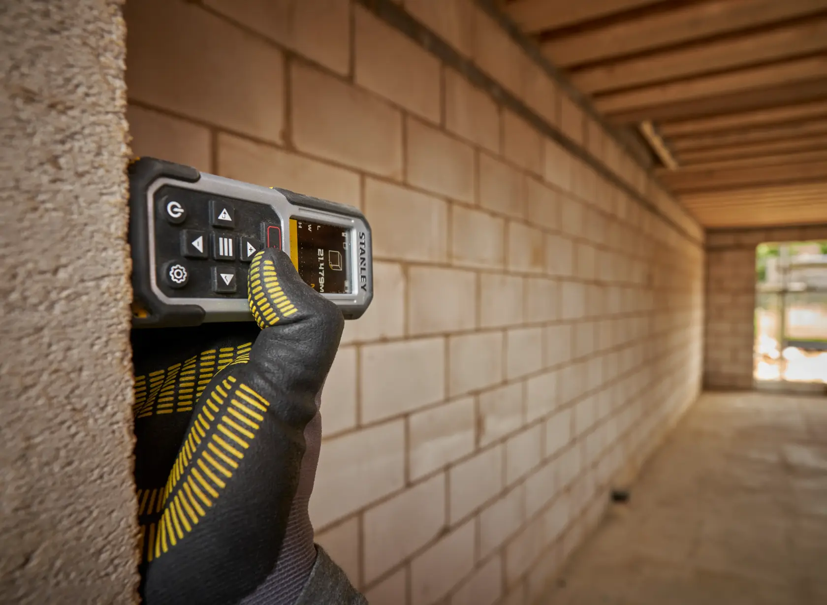 Misuratore laser STANLEY FMHT77050-XJ tenuto da una mano guantata contro un muro di mattoni in cantiere.