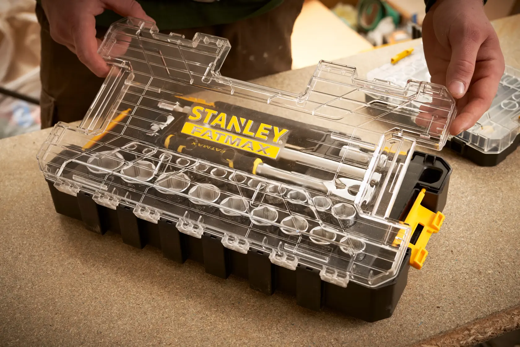 Box organizer STANLEY FMMT98103-1 con coperchio trasparente e FATMAX, tenuto da mani su un banco da lavoro.