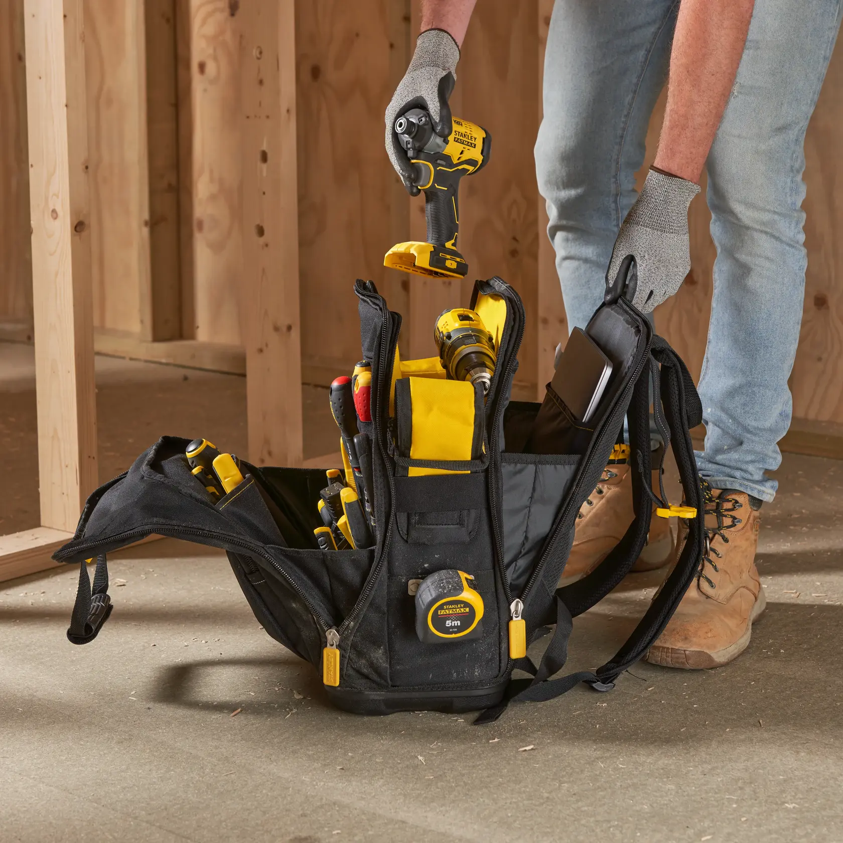 STANLEY FMST1-80144 zaino portautensili con utensili gialli e neri sul pavimento di un cantiere, mani di un lavoratore visibili.