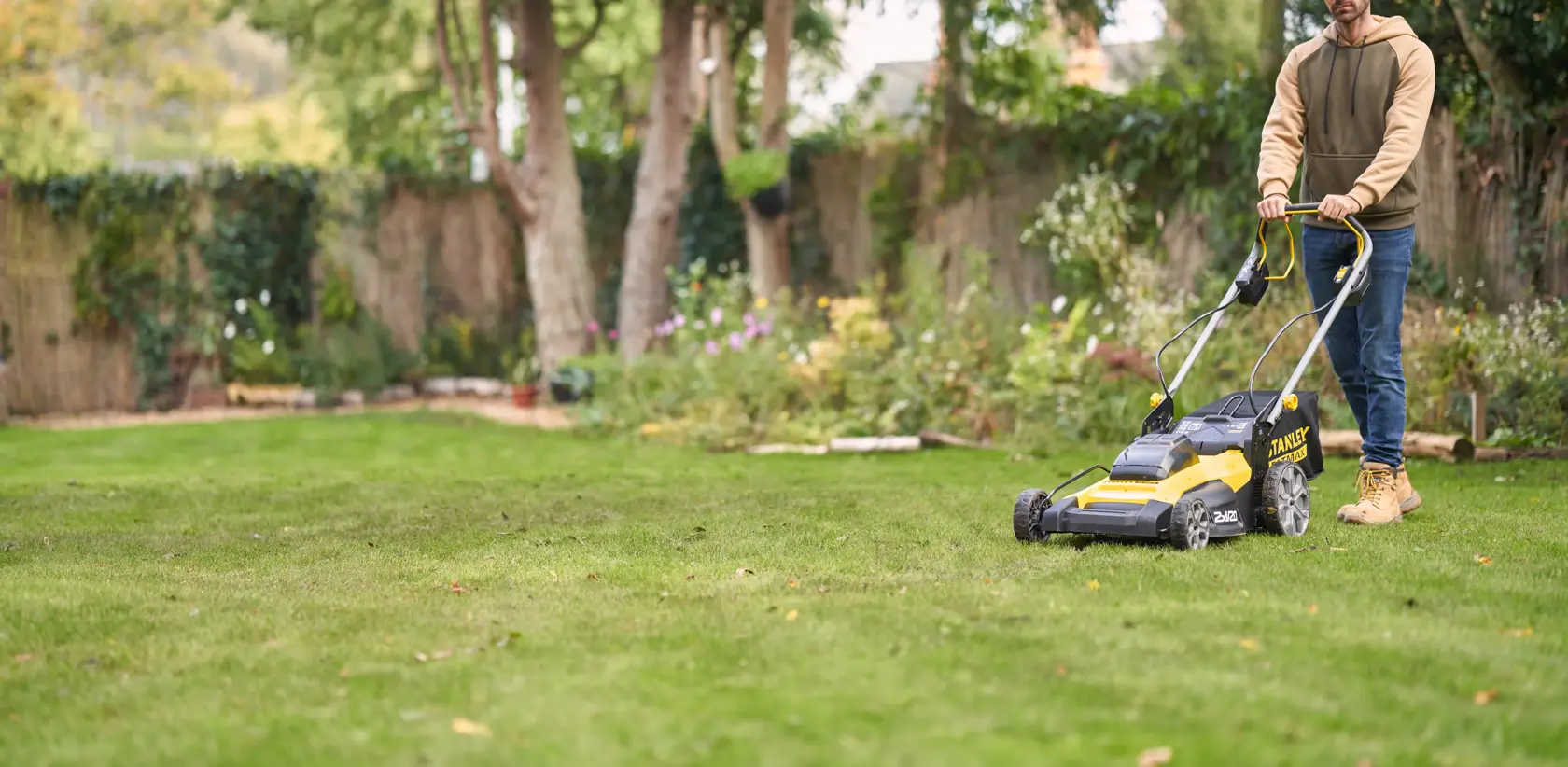 Foto del tosaerba a batteria STANLEY SFMCMWS251B in uso su prato verde, giardino e alberi sullo sfondo.