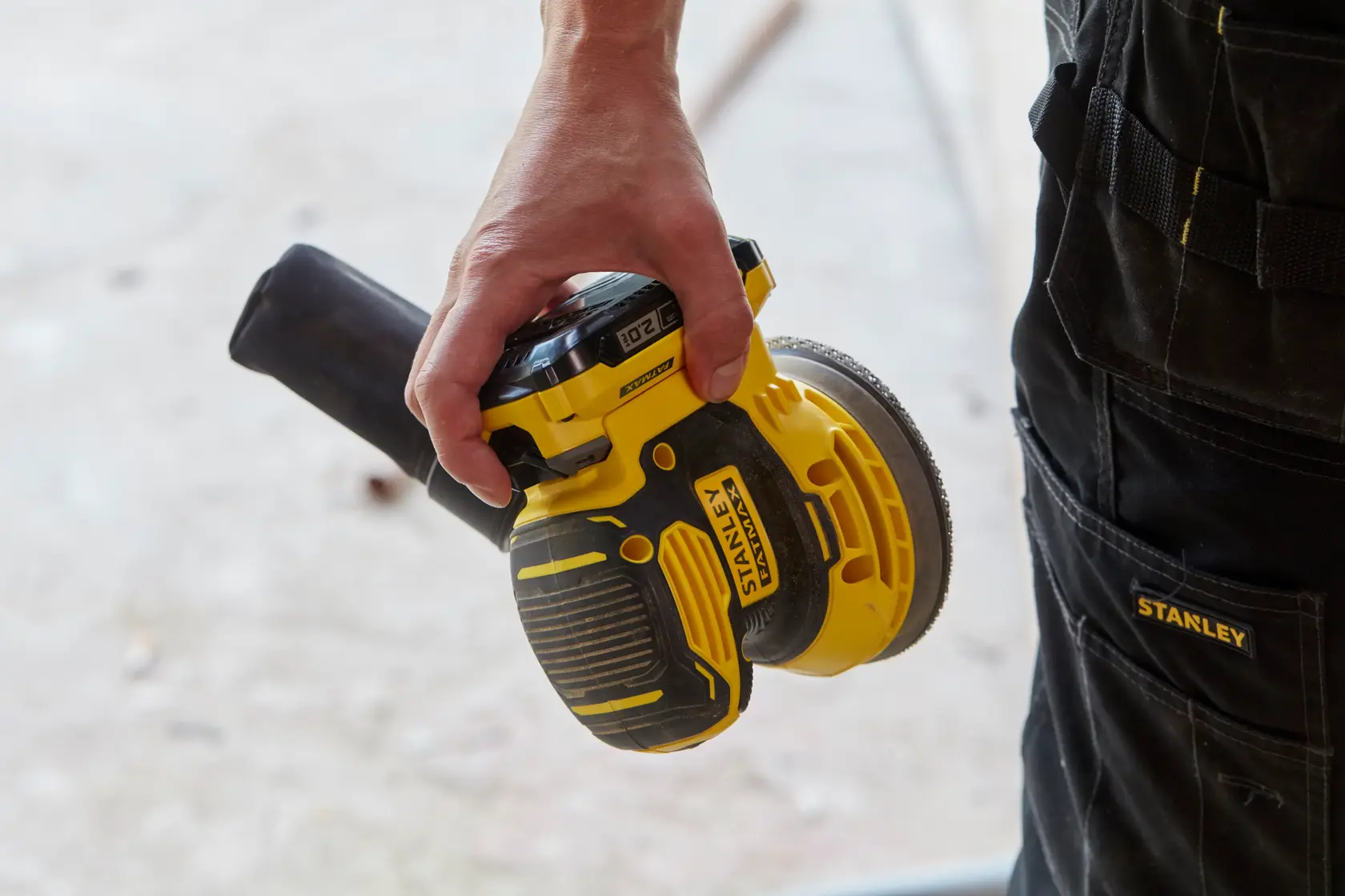 Foto della levigatrice orbitale cordless STANLEY SFMCW220B tenuta in mano, corpo giallo e nero, pantaloni da lavoro visibili.