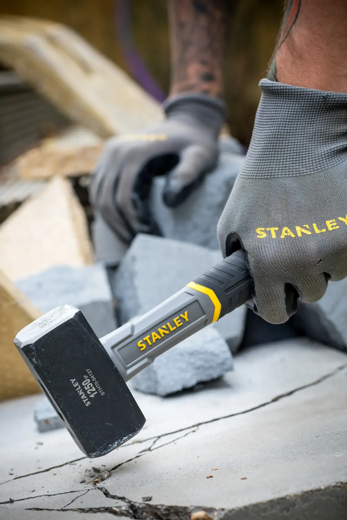 Foto del martello STANLEY STHT0-54127 in uso, testa nera con dettagli gialli, tenuto da mano con guanto su cemento.