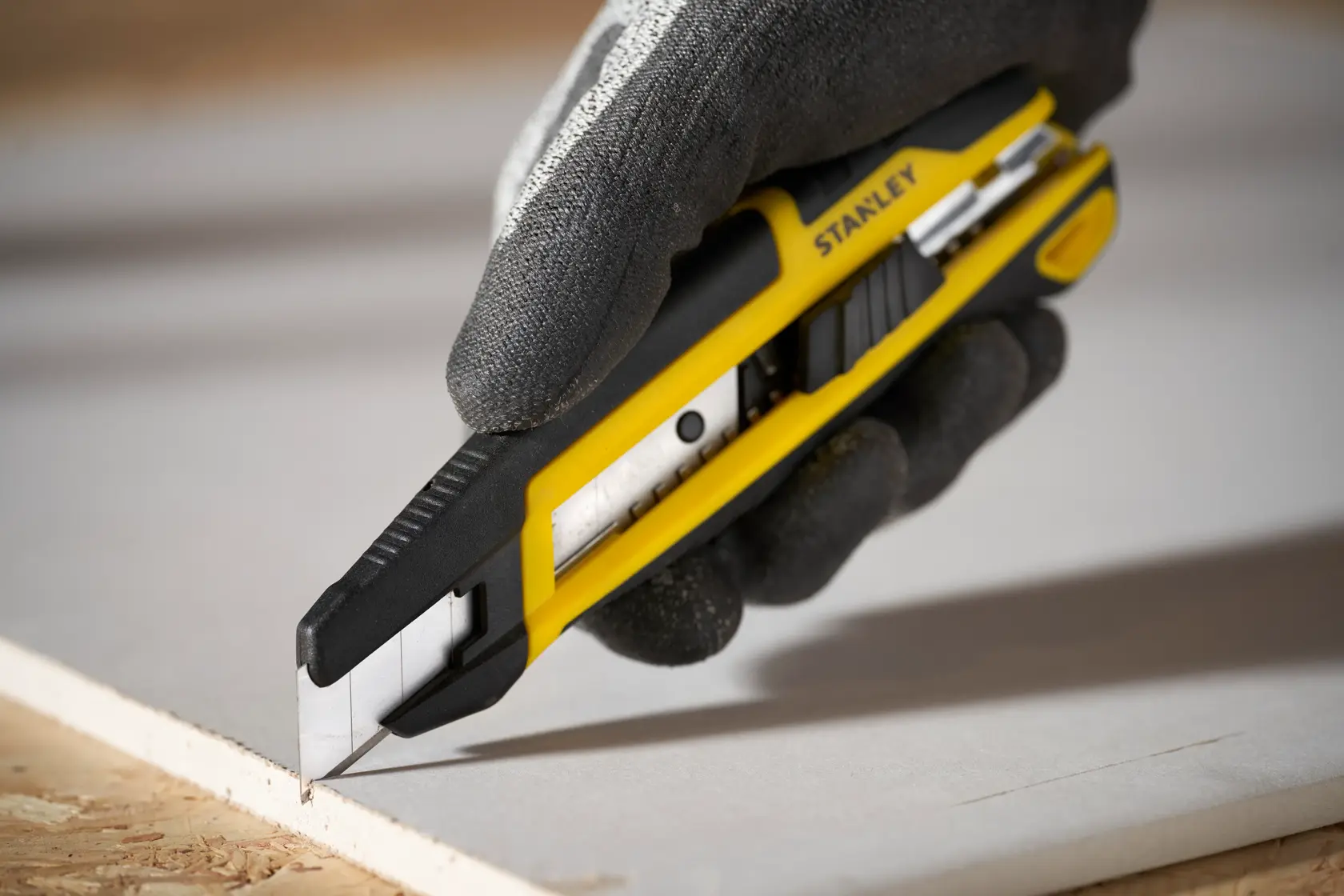Foto del taglierino STANLEY STHT10500-0 giallo e nero che taglia cartongesso, tenuto da una mano guantata.