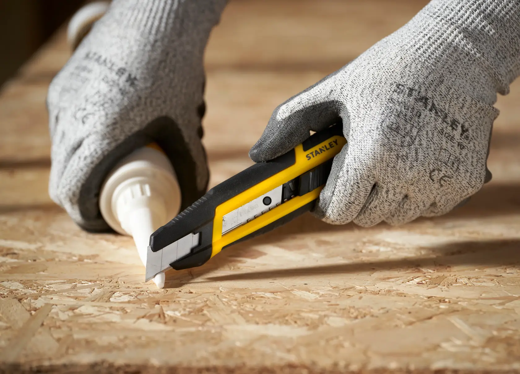 Foto del cutter STANLEY STHT10501-0 che taglia su una superficie in legno, tenuto da mani guantate.