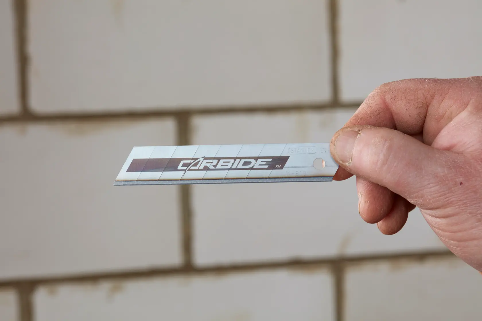 Lama per cutter STANLEY STHT2-11818 argentata con scritta carbide, tenuta in mano davanti a muro piastrellato.