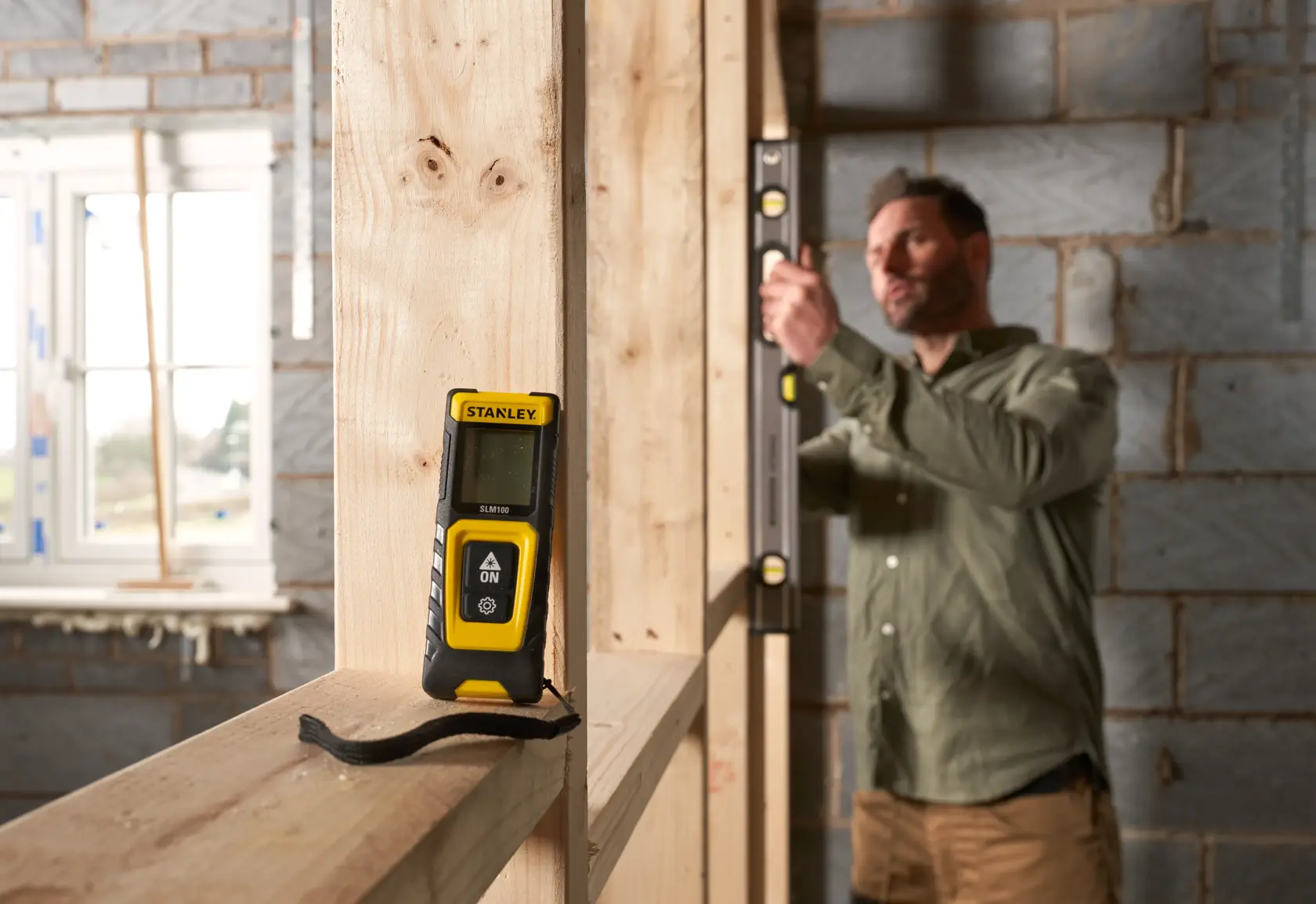 Fotografia del misuratore laser STANLEY STHT77100-0 su telaio in legno in cantiere, con persona che usa una livella sullo sfondo.