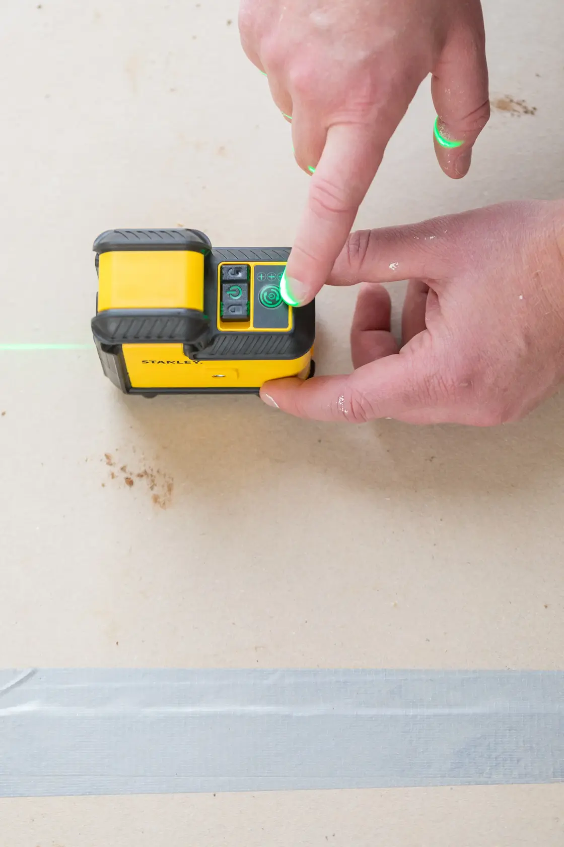 Livella laser STANLEY STHT77594-1 in uso, primo piano della mano che preme il pulsante di accensione, linea laser verde visibile.