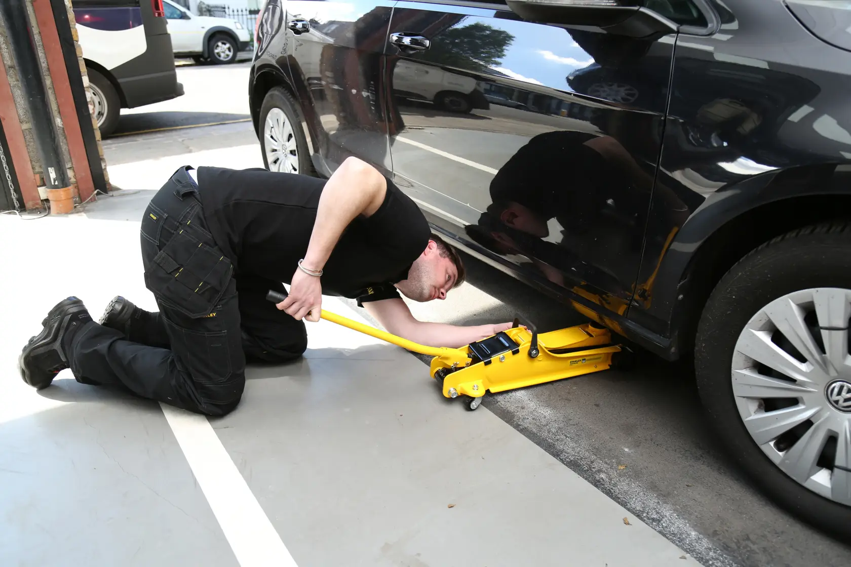 STANLEY STMT81251-1 cric giallo solleva auto nera, tecnico inginocchiato in officina.