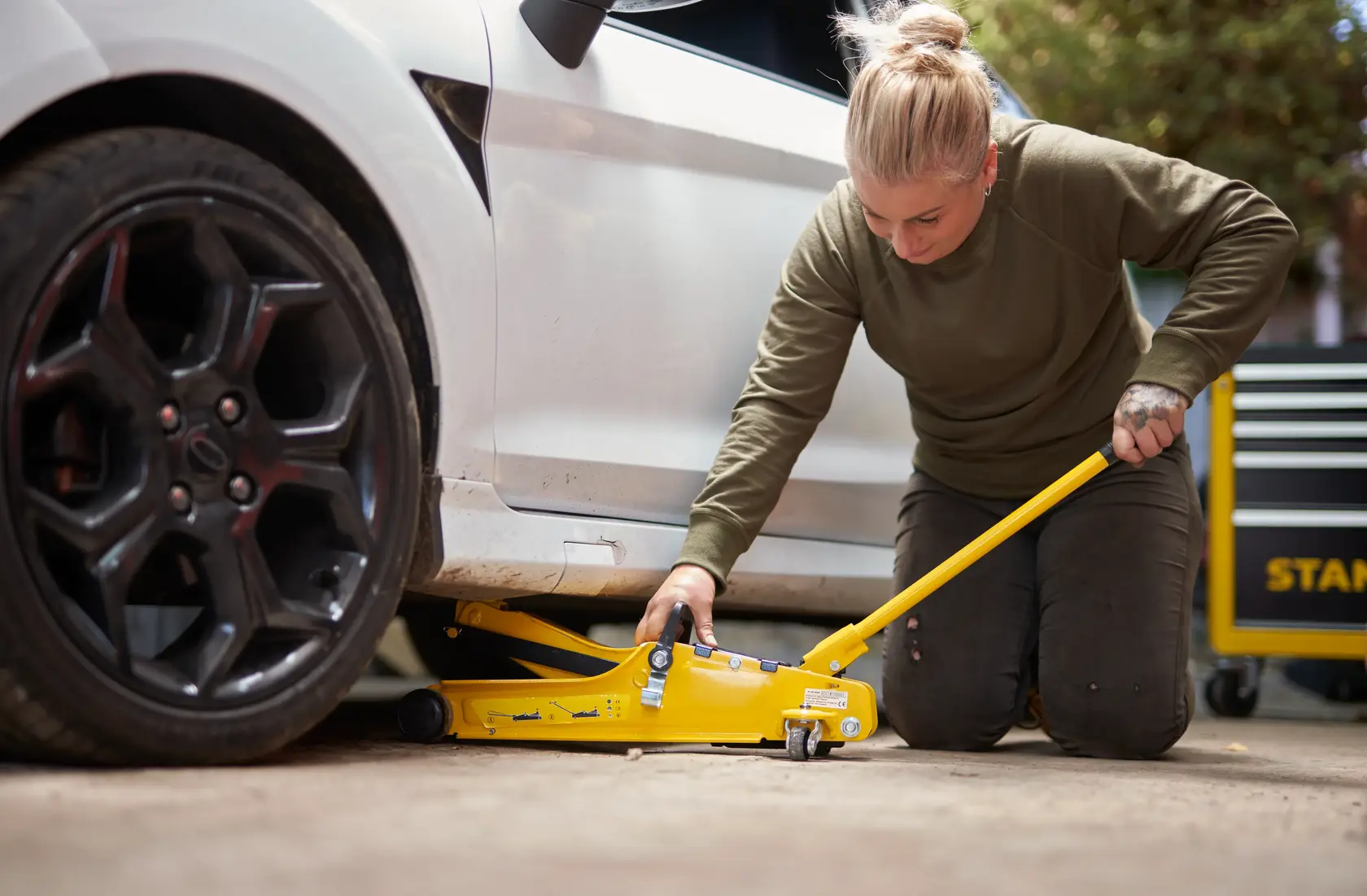 STANLEY STMT81251-1 cric da pavimento giallo in uso solleva un'auto, utente in ginocchio, ambiente garage.