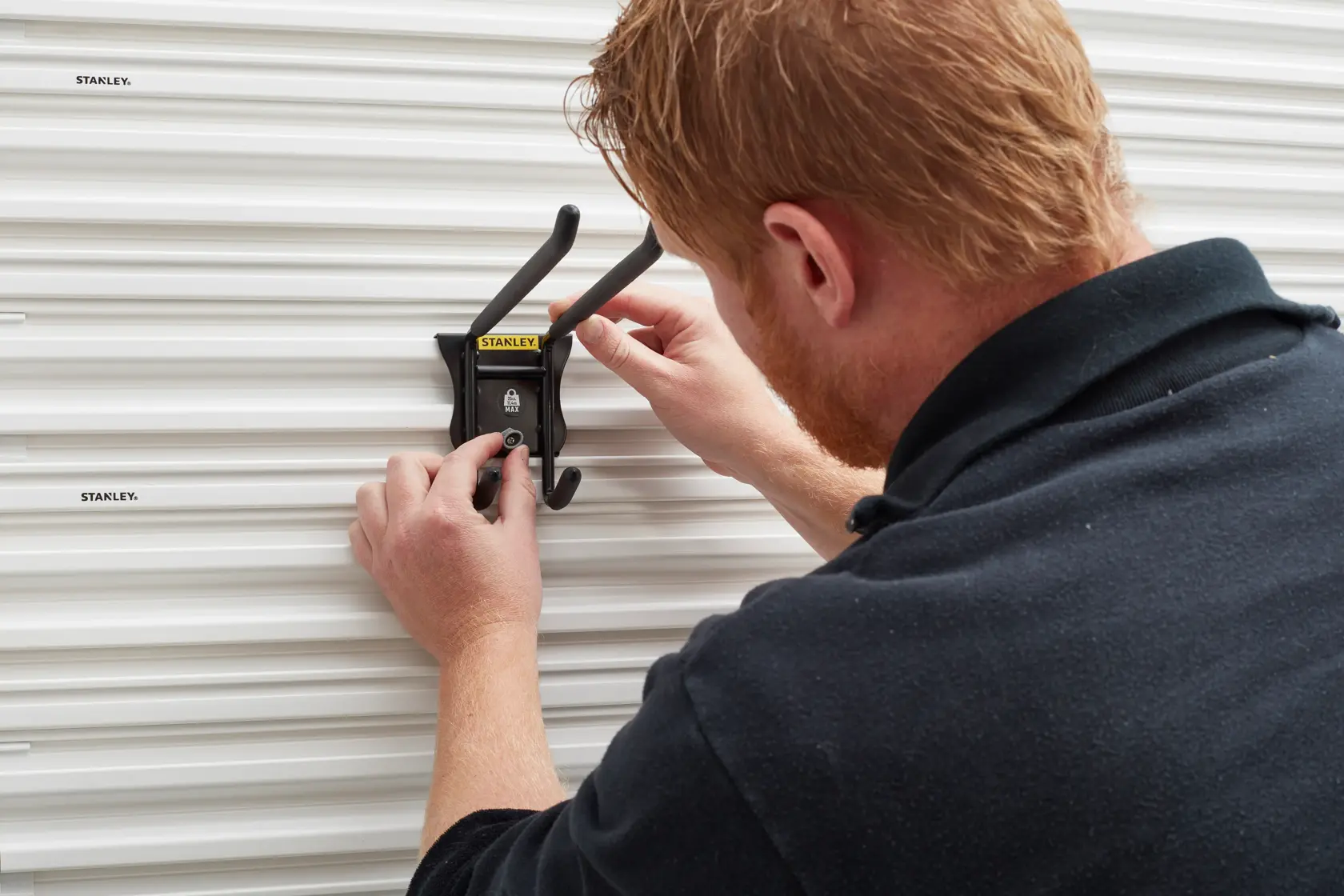 Gancio nero da parete STANLEY STST82606-1 installato su pannello bianco scanalato.